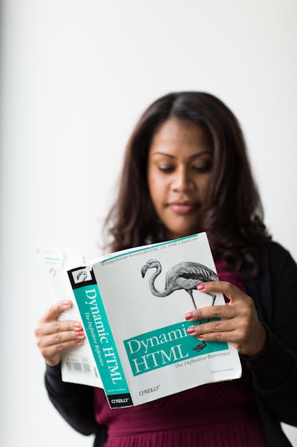 A woman with long dark hair, dressed in a black blazer and dark burgundy top, is holding an open book titled 'Dynamic HTML' with a picture of a flamingo on the cover. The background is plain and white