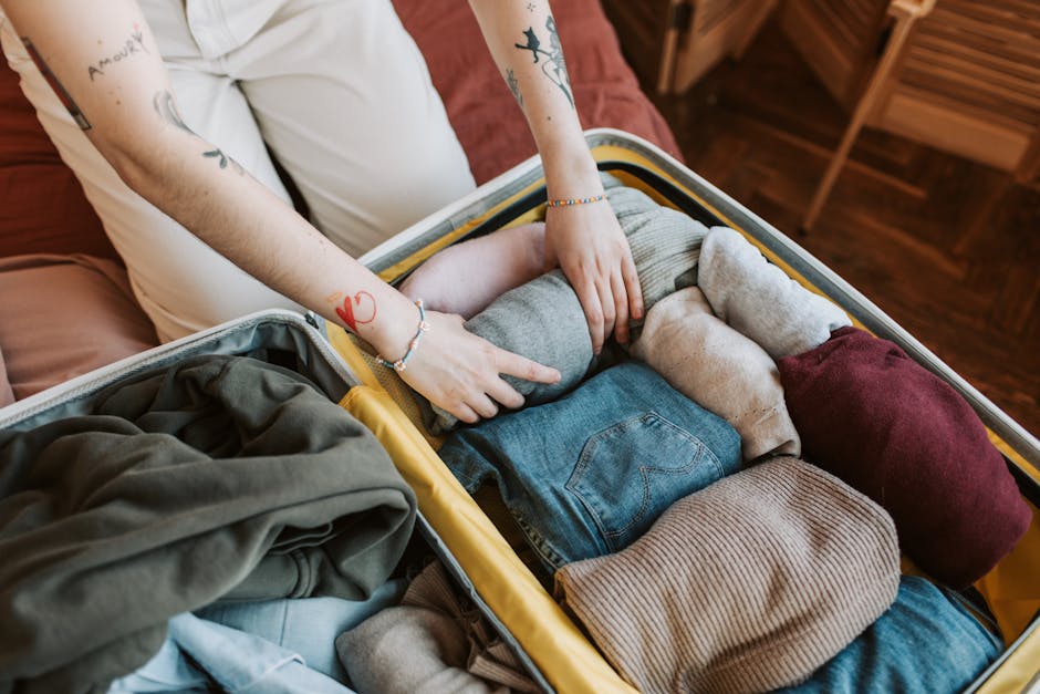 A person with tattoos on both arms is shown packing a black hard-shell suitcase with clothing items such as folded socks, shirts, and a fabric item, all neatly arranged inside. The suitcase is placed